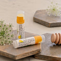 Two tubes of organic beeswax lip balm in Citrus flavor are styled on rustic wooden boards with white flowers and a wooden honey dipper. One tube is standing upright and the other is lying horizontally in the foreground.