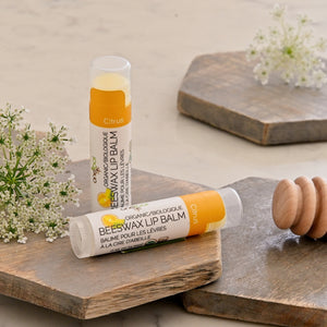 Two tubes of organic beeswax lip balm in Citrus flavor are styled on rustic wooden boards with white flowers and a wooden honey dipper. One tube is standing upright and the other is lying horizontally in the foreground.