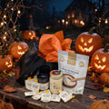 A Halloween-themed gourmet gift set on a rustic wooden table, featuring a black box with orange tissue paper, a bag of organic buckwheat porridge, jars of wildflower and cacao honey, and multiple honey packets, surrounded by glowing jack-o'-lanterns and autumn decorations.